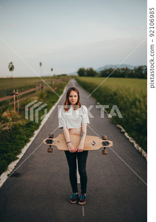 Vertical poster. Pretty woman with tattoos standing in the middle of the road and holding a longboard in her hand 92106985