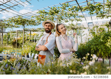 Portrait of colleagues in garden centre. Garden workers in aprons are looking at camera. 92106988