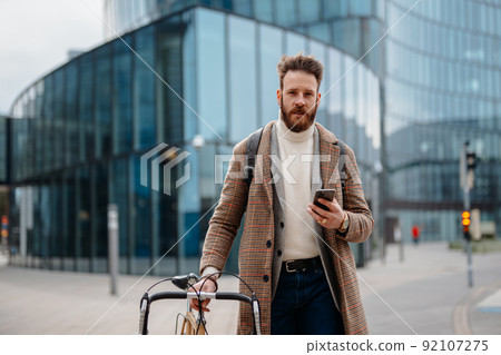 Portrait of hipster businessman with bike, using mobile and looking at camera. Business centre location. Coming to work 92107275