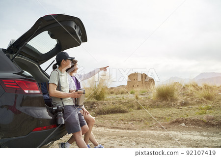 asian couple leaning against back of car looking at view while drinking coffee at desolate historical site asian couple leaning against back of car looking at view while drinking coffee at desolate historical site 92107419
