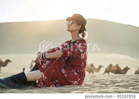 asian woman in red dress sitting in desert looking at view with caravan of camels and huge sand dune in background 92107858
