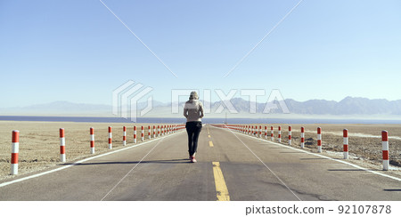 rear view of an asian woman tourist walking on an empty highway towards a lake 92107878