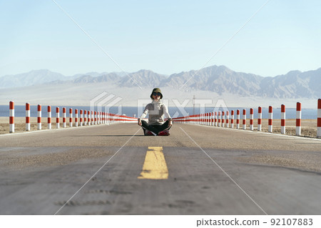 asian woman tourist sitting in the middle of an empty open road with lake and rolling mountains in background, leg crossed. asian woman tourist sitting in the middle of an empty open road with lake and rolling mountains in background, leg crossed. 92107883