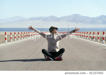 asian woman tourist sitting in the middle of an empty open road with lake and rolling mountains in background, leg crossed arms outstretched 92107885