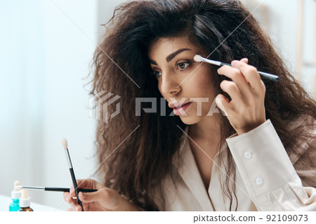 Close up portrait of tanned handsome curly Latin beauty blogger in linen shirt doing makeup in home white interior. Copy space Mockup Banner. Influencer record video review blog using smartphone 92109073