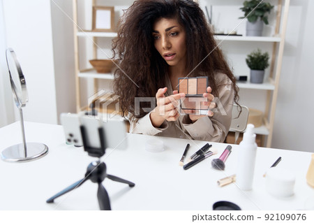 Serious focused tanned handsome curly Latin beauty blogger professional makeup artist in linen shirt record video review blog using smartphone in home white interior. Copy space. Influencer concept Serious focused tanned handsome curly Latin beauty blogger professional makeup artist in linen shirt record video review blog using smartphone in home white interior. Copy space. Influencer concept 92109076