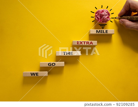 We go the extra mile symbol. Wood blocks stacking as step stair on beautiful yellow background, copy space. Male hand, light bulb. Words 'We go the extra mile'. Business and go the extra mile concept. We go the extra mile symbol. Wood blocks stacking as step stair on beautiful yellow background, copy space. Male hand, light bulb. Words 'We go the extra mile'. Business and go the extra mile concept. 92109285