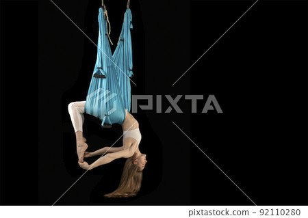 Young woman in white sportswear practices aerial inversion anti-gravity yoga with hammock isolated on black background. Side view. 92110280