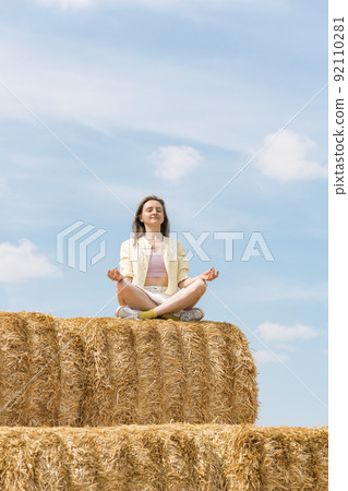 Happy young woman sits in lotus position on huge haystack and meditates. Blue sky background. Countryside holiday. 92110281