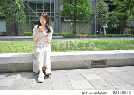 A young woman sitting on a bench and operating a smartphone 92118668