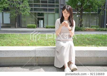 A young woman sitting on a bench and operating a smartphone 92118669
