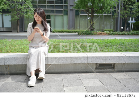 A young woman sitting on a bench and operating a smartphone 92118672