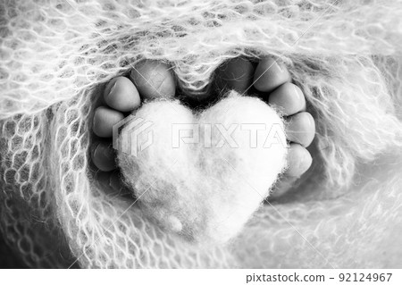 The tiny foot of a newborn baby. Soft feet of a new born in a wool blanket. Close up of toes, heels and feet of a newborn. Knitted heart in the legs of baby. Macro photography. Black and white. The tiny foot of a newborn baby. Soft feet of a new born in a wool blanket. Close up of toes, heels and feet of a newborn. Knitted heart in the legs of baby. Macro photography. Black and white. 92124967