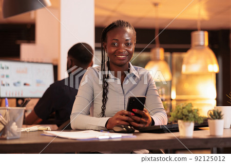 Portrait of executive manager using mobile phone with touchscreen, browsing social media app and sitting at office desk. Working with computer and smartphone to do research report. 92125012