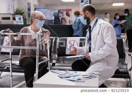 Elderly woman with impairment in waiting room lobby doing checkup consultation with specialist during coronavirus pandemic. Patient with walk frame having appointment in hospital reception. Elderly woman with impairment in waiting room lobby doing checkup consultation with specialist during coronavirus pandemic. Patient with walk frame having appointment in hospital reception. 92125014