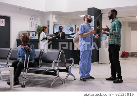 African american man talking to medical assistant in ospital reception lobby, discussing about disease diagnosis and recovery treatment. Busy waiting room with sick patients at clinic. African american man talking to medical assistant in ospital reception lobby, discussing about disease diagnosis and recovery treatment. Busy waiting room with sick patients at clinic. 92125019
