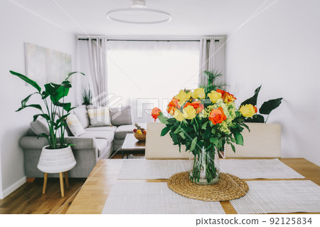 Kitchen counter table with focus on vase with yellow and orange roses on it with blurred background of modern cozy living room with couch and green plants. Open space home interior design. Copy space. Kitchen counter table with focus on vase with yellow and orange roses on it with blurred background of modern cozy living room with couch and green plants. Open space home interior design. Copy space. 92125834