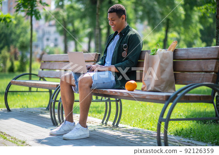 Guy working on laptop sitting on bench outdoors Guy working on laptop sitting on bench outdoors 92126359