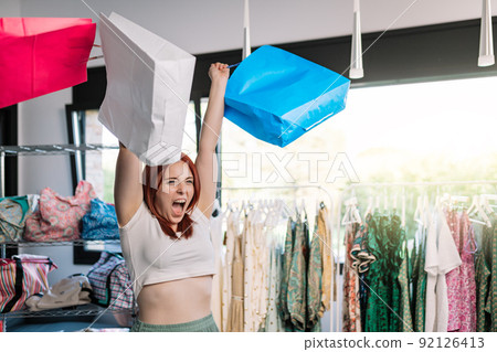 young red-haired woman shouting with joy, raising her hands and holding paper bags, shopping in a clothing shop. concept of shopping. young red-haired woman shouting with joy, raising her hands and holding paper bags, shopping in a clothing shop. concept of shopping. 92126413