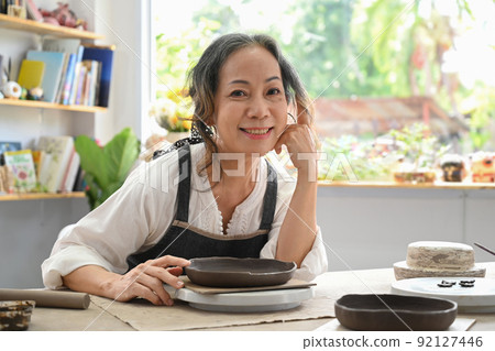 Smiling retired woman creating handicraft crockery in pottery workshop. Activity, handicraft, hobbies concept Smiling retired woman creating handicraft crockery in pottery workshop. Activity, handicraft, hobbies concept 92127446