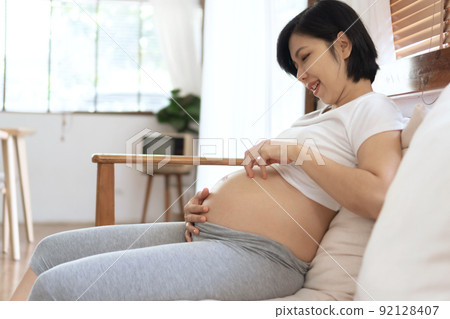 Chinese or Japanese Female playing her pregnant belly sitting on sofa 92128407