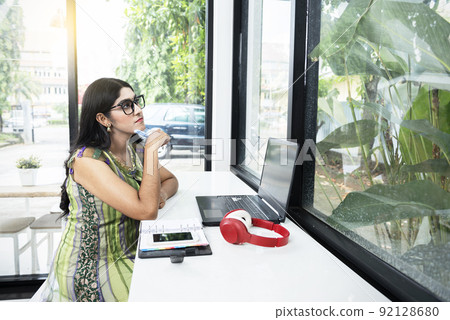 Indian woman with eyeglass using a laptop with a notebook and a mobile phone on the table Indian woman with eyeglass using a laptop with a notebook and a mobile phone on the table 92128680