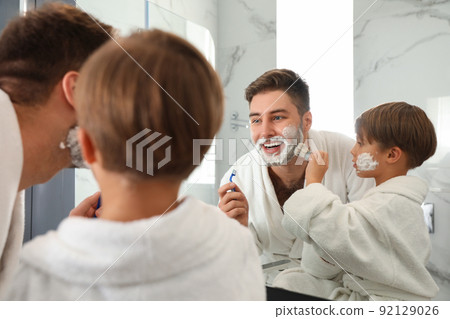 Dad and son with shaving foam on their faces having fun in bathroom Dad and son with shaving foam on their faces having fun in bathroom 92129026