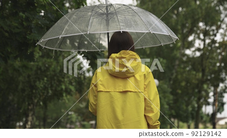 a young girl with a transparent umbrella walks in the rain. woman in a yellow raincoat. view from the back. autumn summer spring walk park. Slow motion. wet drops rain drip transparent umbrella. a young girl with a transparent umbrella walks in the rain. woman in a yellow raincoat. view from the back. autumn summer spring walk park. Slow motion. wet drops rain drip transparent umbrella. 92129142