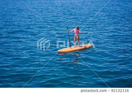 Young women Having Fun Stand Up Paddling in blue water sea in Montenegro. SUP. girl Training on Paddle Board near the rocks Young women Having Fun Stand Up Paddling in blue water sea in Montenegro. SUP. girl Training on Paddle Board near the rocks 92132116