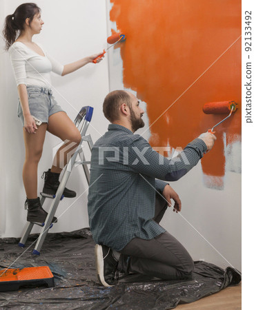 Young couple using orange paint to renovate apartment walls and change house design, painting with vibrant color using paintbrush, roller and tools. Diy home renovation and decoration. Young couple using orange paint to renovate apartment walls and change house design, painting with vibrant color using paintbrush, roller and tools. Diy home renovation and decoration. 92133492