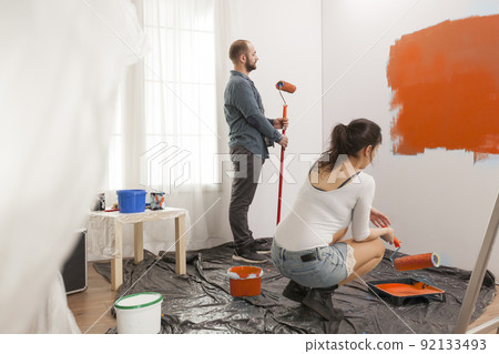 Family painting room with orange color paint to redecorate apartment with paintbrush and roller. Couple doing diy manual renovation using decorating tools and equipment, housework change. Family painting room with orange color paint to redecorate apartment with paintbrush and roller. Couple doing diy manual renovation using decorating tools and equipment, housework change. 92133493