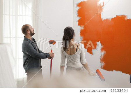 Man and woman painting with orange color, using paint to redecorate walls in apartment. Renovating house interior with paintbrush, renovation tools and roller, decorating room. Man and woman painting with orange color, using paint to redecorate walls in apartment. Renovating house interior with paintbrush, renovation tools and roller, decorating room. 92133494