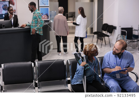 Health specialist consulting woman with crutches in hospital reception lobby, talking about diagnosis and medicine in waiting room area. Diverse people discussing at checkup appointment. 92133495