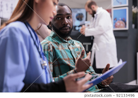Asian assistant explaining diagnosis to male patient in hospital reception, doing checkup consultation in waiting area. Young man and specialist talking about healthcare treatment at clinic. Asian assistant explaining diagnosis to male patient in hospital reception, doing checkup consultation in waiting area. Young man and specialist talking about healthcare treatment at clinic. 92133496