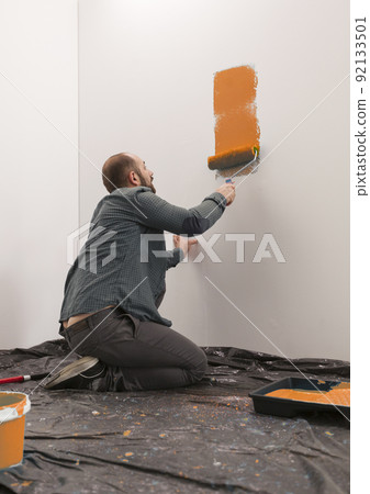 Man painting wall with orange paint, using paintbrush and roller to redecorate apartment room. Renovating home with diy tools and equipment. Vibrant paint renovation and improvement. Man painting wall with orange paint, using paintbrush and roller to redecorate apartment room. Renovating home with diy tools and equipment. Vibrant paint renovation and improvement. 92133501