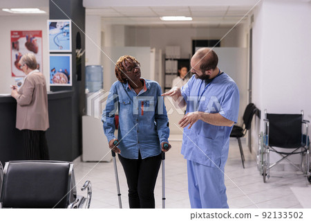 Female patient with crutches talking to nurse in medical facility lobby, waiting to attend healthcare consultation with specialist. Woman dealing with physical impairment at hospital reception. 92133502