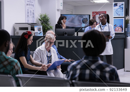 Senior pediatrician with clipboard doctor completing form for mother and child for routine checkup appointment in hospital reception. Diverse people waiting in modern private practice clinic. 92133649