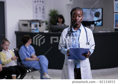 Portrait of african american doctor with stethoscope looking at clipboard with patient data in hospital reception. Confident medic in clinic reception lobby waiting for patient appointment. 92133653