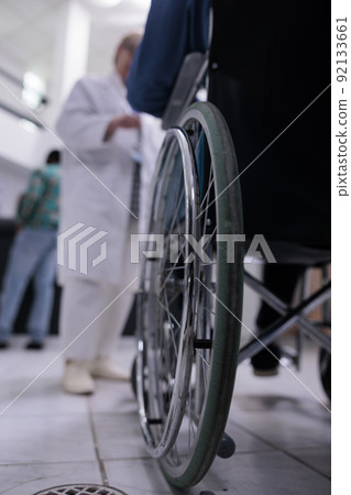 Closeup of senior man living with disability using wheelchair in private clinic reception talking with medical doctor about appointment. Selective focus on wheelchair wheel in busy hospital lobby. 92133661