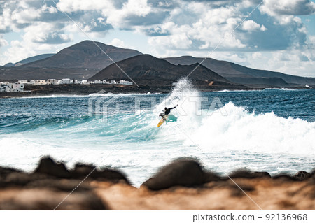 Wild rocky coastline of surf spot La Santa Lanzarote, Canary Islands, Spain. Surfer riding a big wave in rocky bay, volcano mountain in background. 92136968