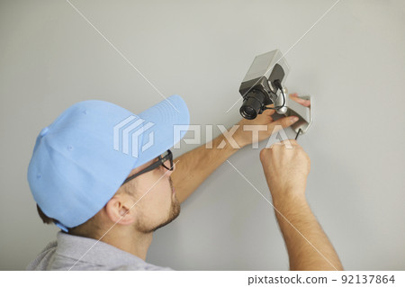 Young male worker installing surveillance CCTV camera on wall inside the building Young male worker installing surveillance CCTV camera on wall inside the building 92137864