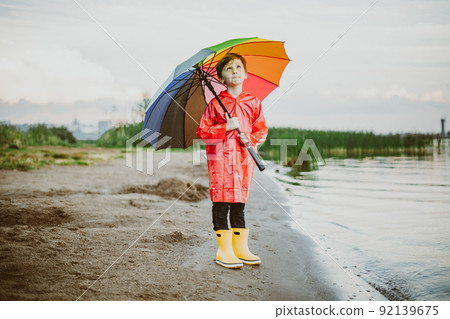 Boy in a red raincoat and yellow rubber boots stands at river bank and holding rainbow umbrella. School kid standing still near autumn lake. Child wearing waterproof clothes at shoreside 92139675