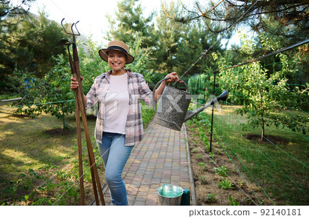 Charming female gardener poses to camera with gardening tools- watering can and hoe against an organic farm background Charming female gardener poses to camera with gardening tools- watering can and hoe against an organic farm background 92140181