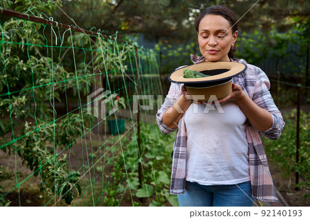 Female gardener amateur smelling freshly harvested ripe cucumbers in straw hat while working in an ecological farm. Female gardener amateur smelling freshly harvested ripe cucumbers in straw hat while working in an ecological farm. 92140193