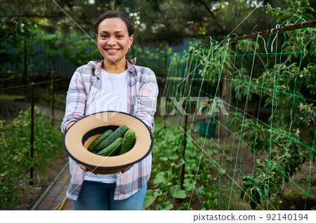 Beautiful woman gardener smiling toothy smile, holding a straw hat with freshly picked ripe cucumbers in organic farm Beautiful woman gardener smiling toothy smile, holding a straw hat with freshly picked ripe cucumbers in organic farm 92140194