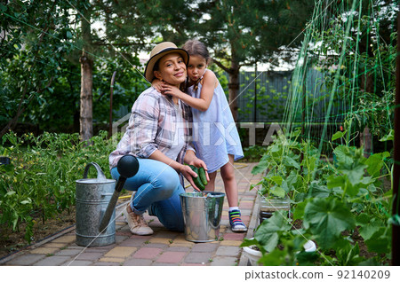Adorable little girl hugs her mother while picking ripe cucumbers in the country house eco garden. Family agribusiness.  92140209