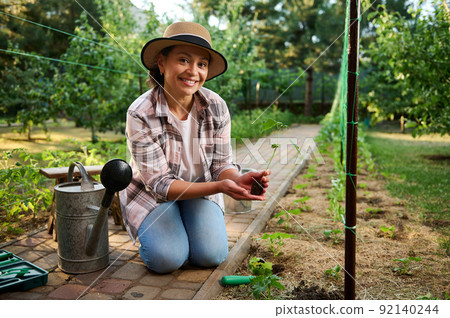 Caucasian pretty woman, farmer sitting near a watering can, holding a seedling of cucumbers, smiling looking at camera.  92140244