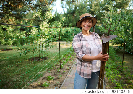 Caucasian woman, farmer, horticulturist, gardener smiles to the camera with gardening tools in her hands in organic farm  92140266