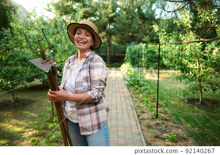 Cheerful woman, gardener holding garden tools in her hands standing in rows of planted seedlings on an early spring day  92140267