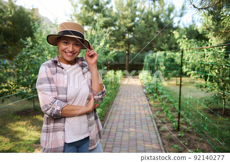 Pleasant pretty Hispanic woman, female gardener smiles looking at camera, against an organic vegetable garden background  92140277